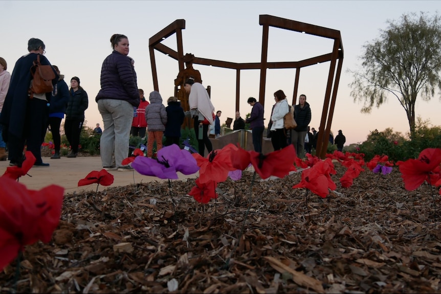 A group of people stand near a pole monument with Poppy flowers at the forefront. 
