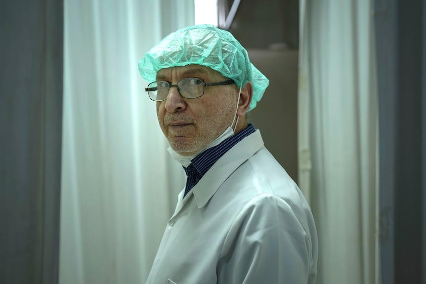 A man wearing a white jacket, glasses and green cap stands staring at the camera.