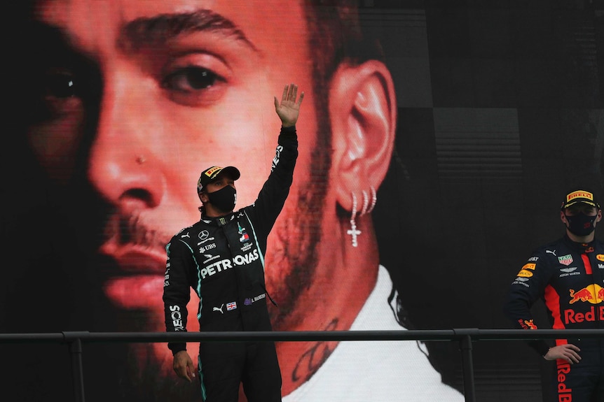 Standing in front of a giant picture of his own face, Lewis Hamilton waves to the crowd.