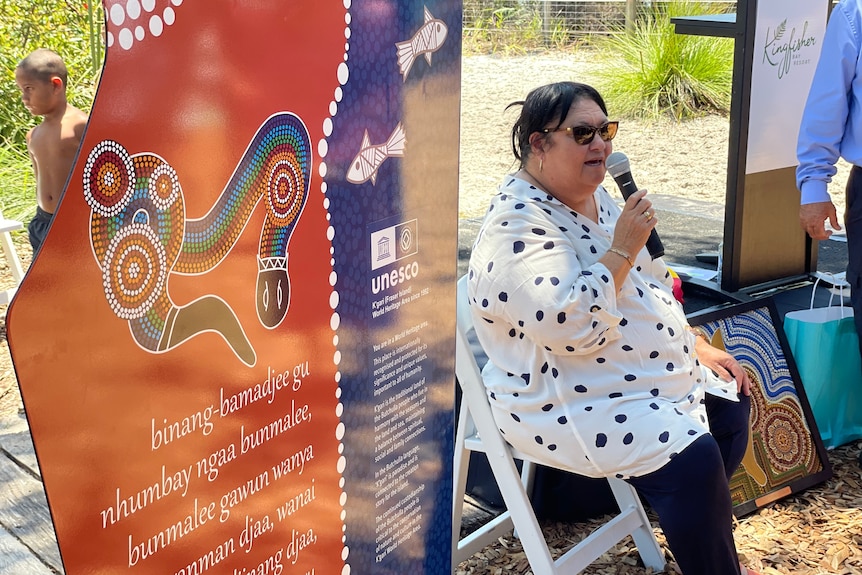 A large woman, wears white polka dot top, black pants holds microphone sits in a chair on the beach,  Aboriginal art beside her.