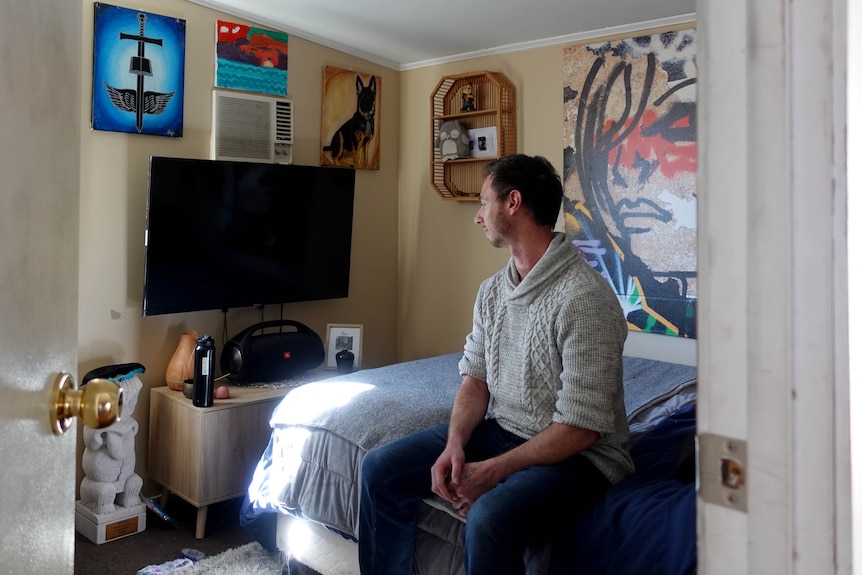 A man in a grey jumper sits on a bed in a room with a tv, and paintings on the wall. 
