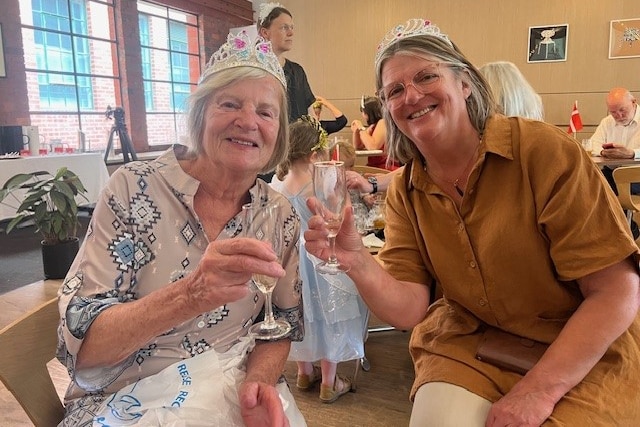 Two women wearing tiaras hold glasses of champagne and smile at the camera