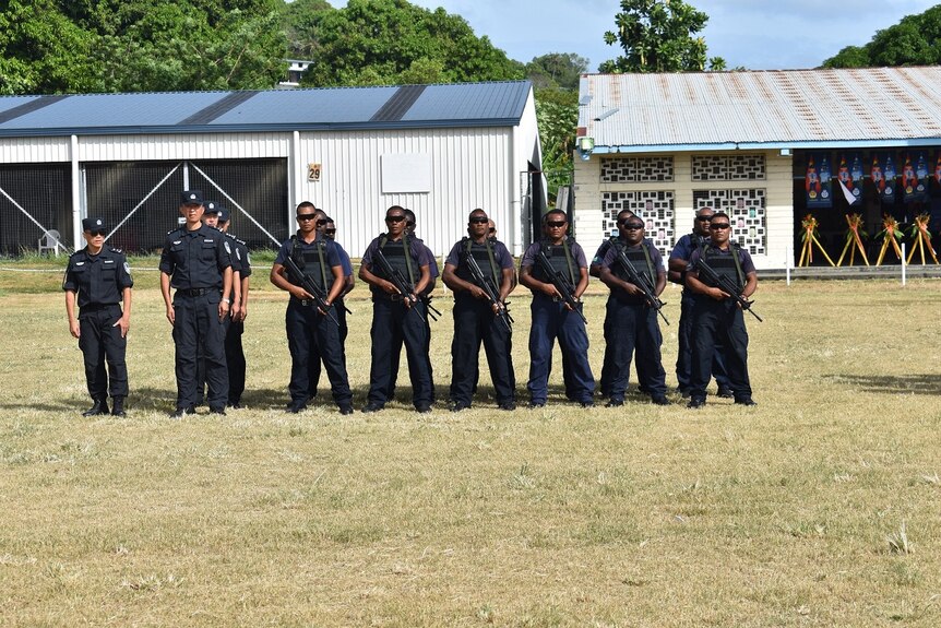 Police stand in formation in navy uniforms with rifles pointed at the grass.