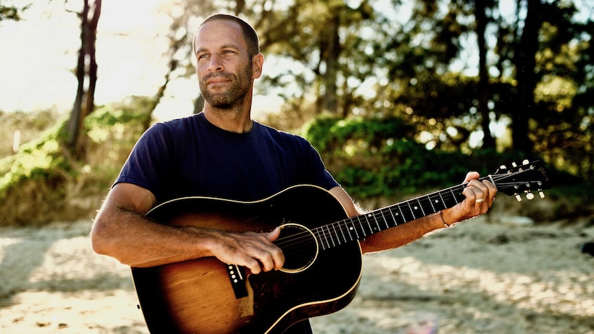 Jack Johnson stands on a beach playing an acoustic guitar