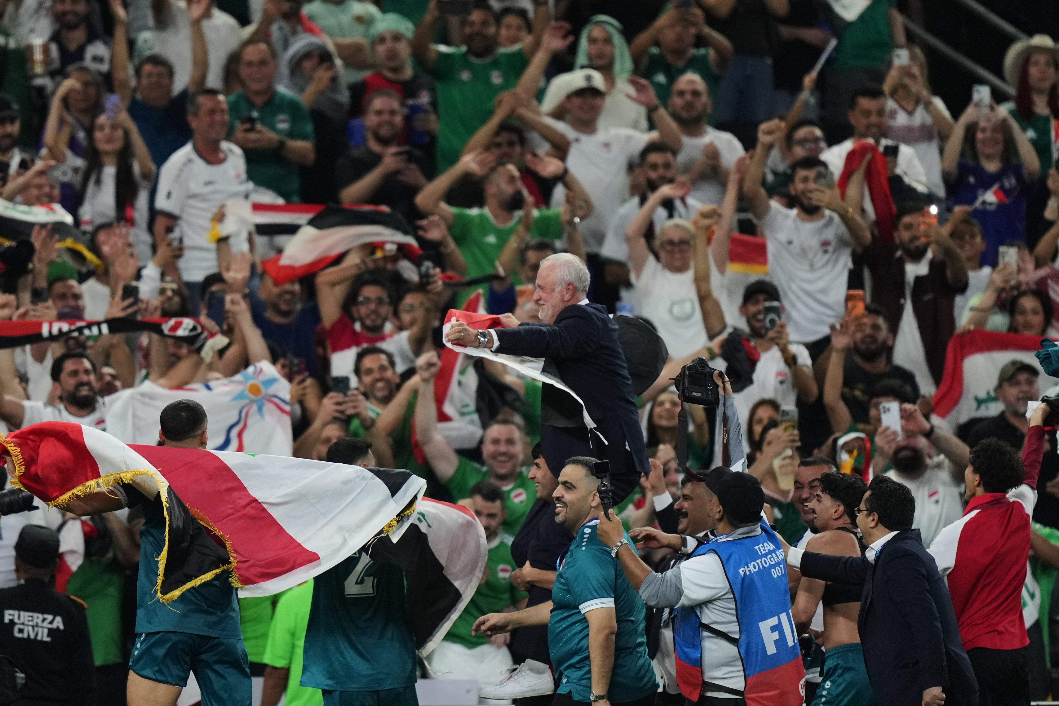 Male footballers carry an older man in a suit, who waves an Iraqi flag, off a soccer field in front of a huge, ecstatic crowd.