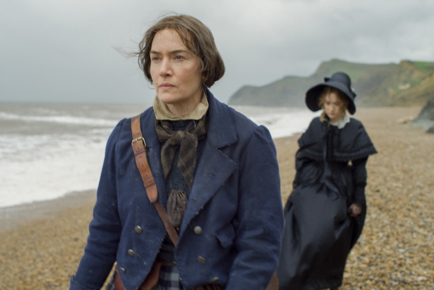 Winslet and Ronan in 19th century clothing walking on overcast beach towards camera, Winslet leading and Ronan lagging behind.