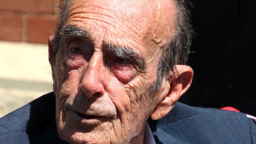 An elderly man in a suit jacket outside a brick building.