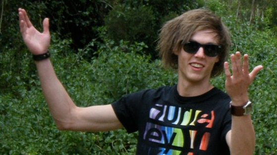 Young man with medium length brown hair and sunglasses smiling with his arms up.