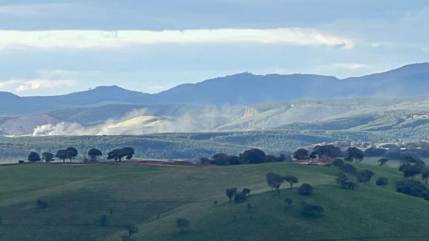 Plumes of dust rising off the site of a mine with hills in the foreground and background 