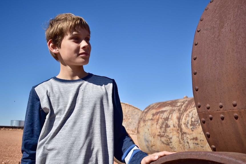 A young boy stands next to a rusted metal farming implement against a blue sky