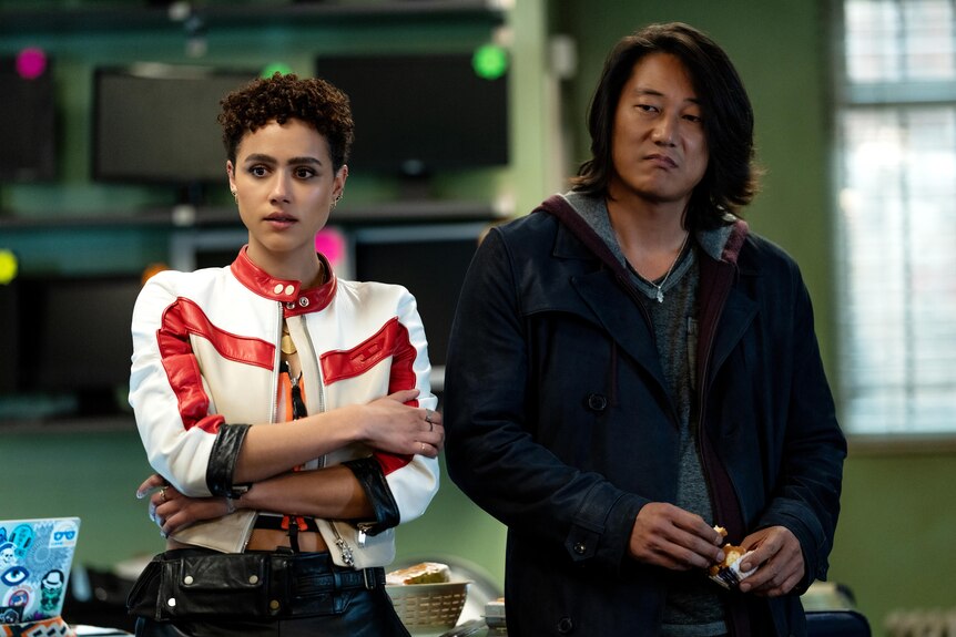 A fair-skinned black woman with curly hair wearing a racing jacket and a long-haired Asian man in a black hoodie stand together.