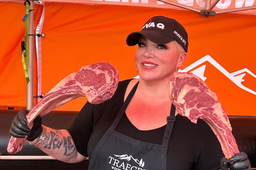 A woman wearing a black hat and apron is holding two large pieces of meat
