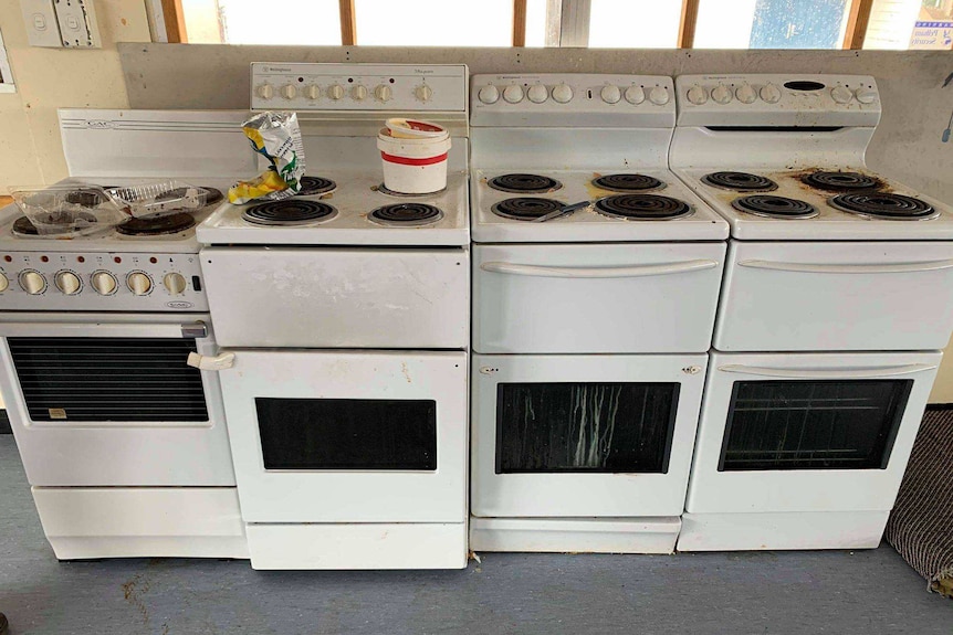 Four stoves side-by-side in a kitchen. They are untidy and have rubbish on them
