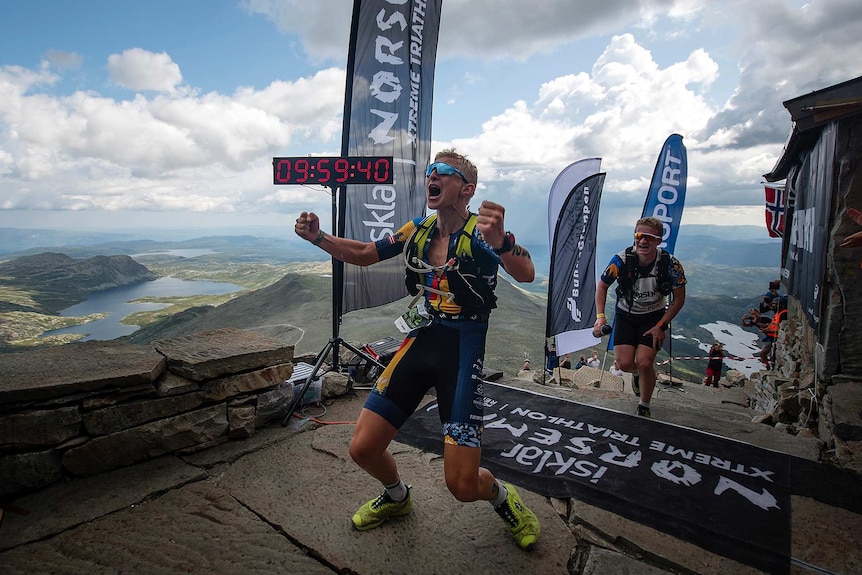 A triathlon runner screams in delight after crossing the finish line with mountains in the background