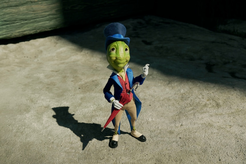 Live-action still of humanoid cricket wearing a blue cloak and top hat with red umbrella and vest.