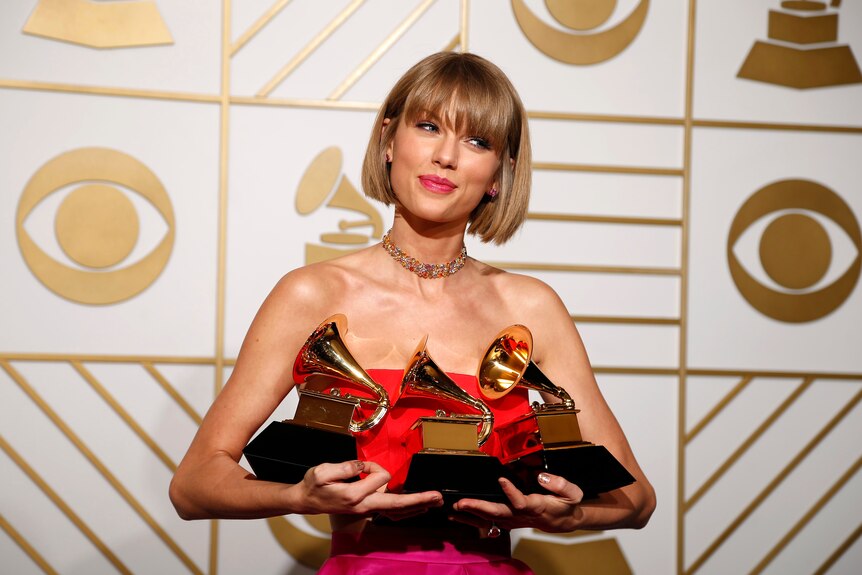 Taylor smiles softly and tilts her head to the side as she holds up three Grammy Awards.