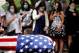 A group of women in face masks with their hands on their hearts behind a coffin draped in a US flag