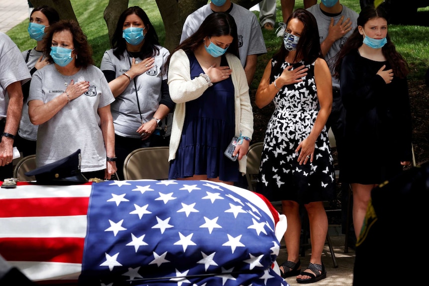 A group of women in face masks with their hands on their hearts behind a coffin draped in a US flag