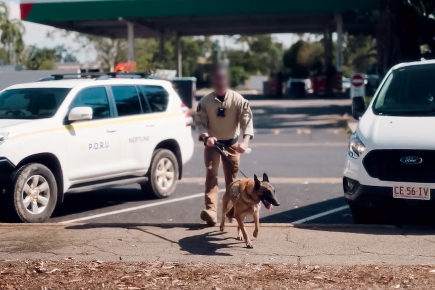 A man in tan/khaki uniform and sunglasses steps onto a footpath towards the camera with a guard dog on a lead.
