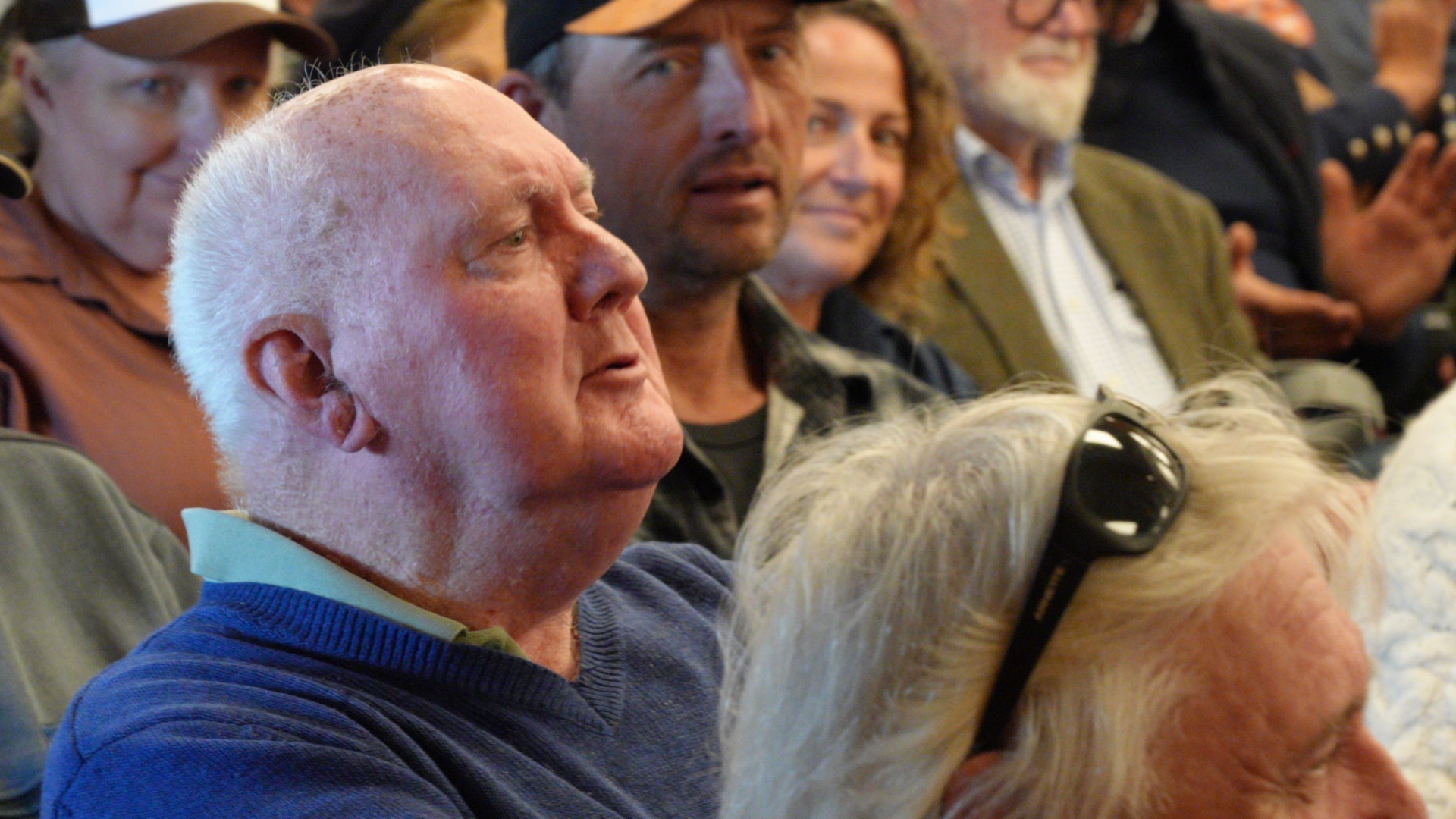 An older man sits among a crowd in a community hall.