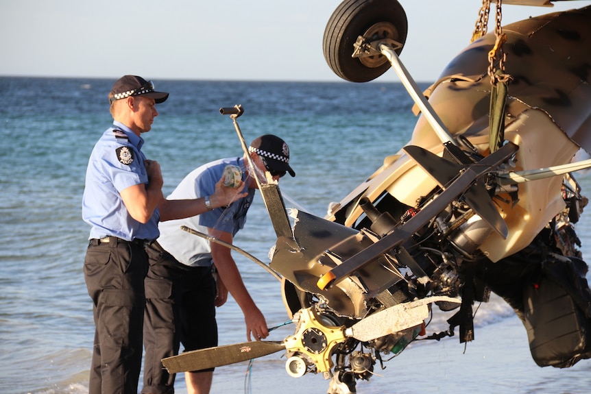 Two policemen examine the wreckage of a gyrocopter suspended upside down from a chain on a beach