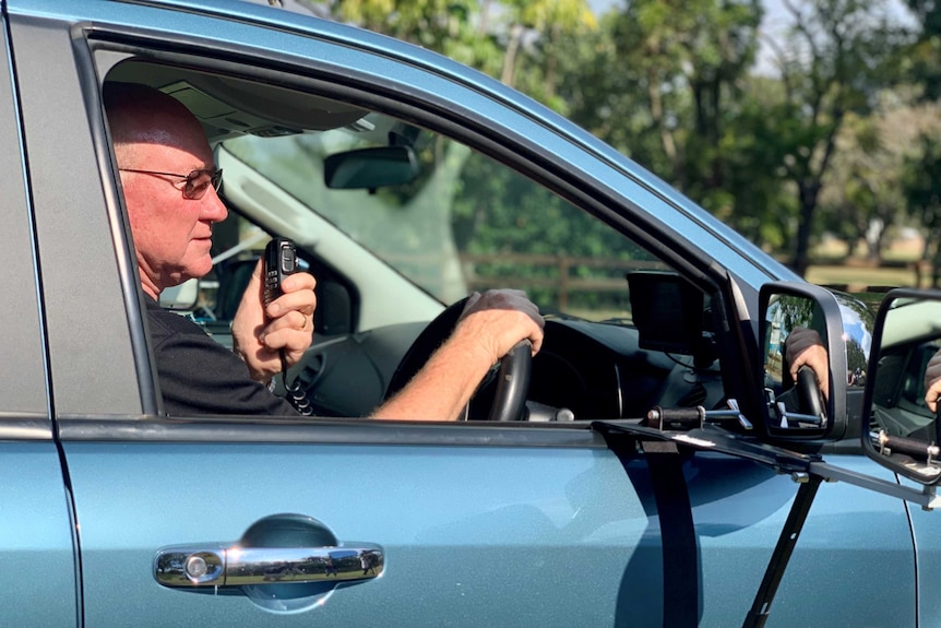 A man sits in a blue car that is towing a caravan, while talking into a UHF radio