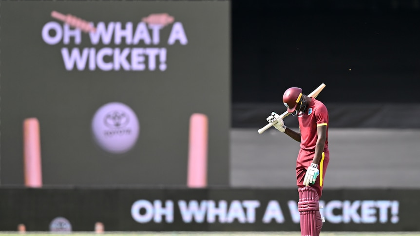 A dejected West Indies batter looks down at his feet while a big screen behind him shows a sign "Oh what a wicket!". 