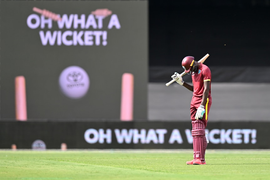 A dejected West Indies batter looks down at his feet while a big screen behind him shows a sign "Oh what a wicket!". 