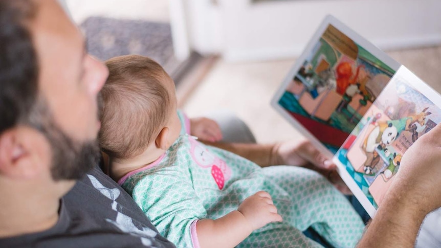 A man reads a children's book to a baby in his arms.
