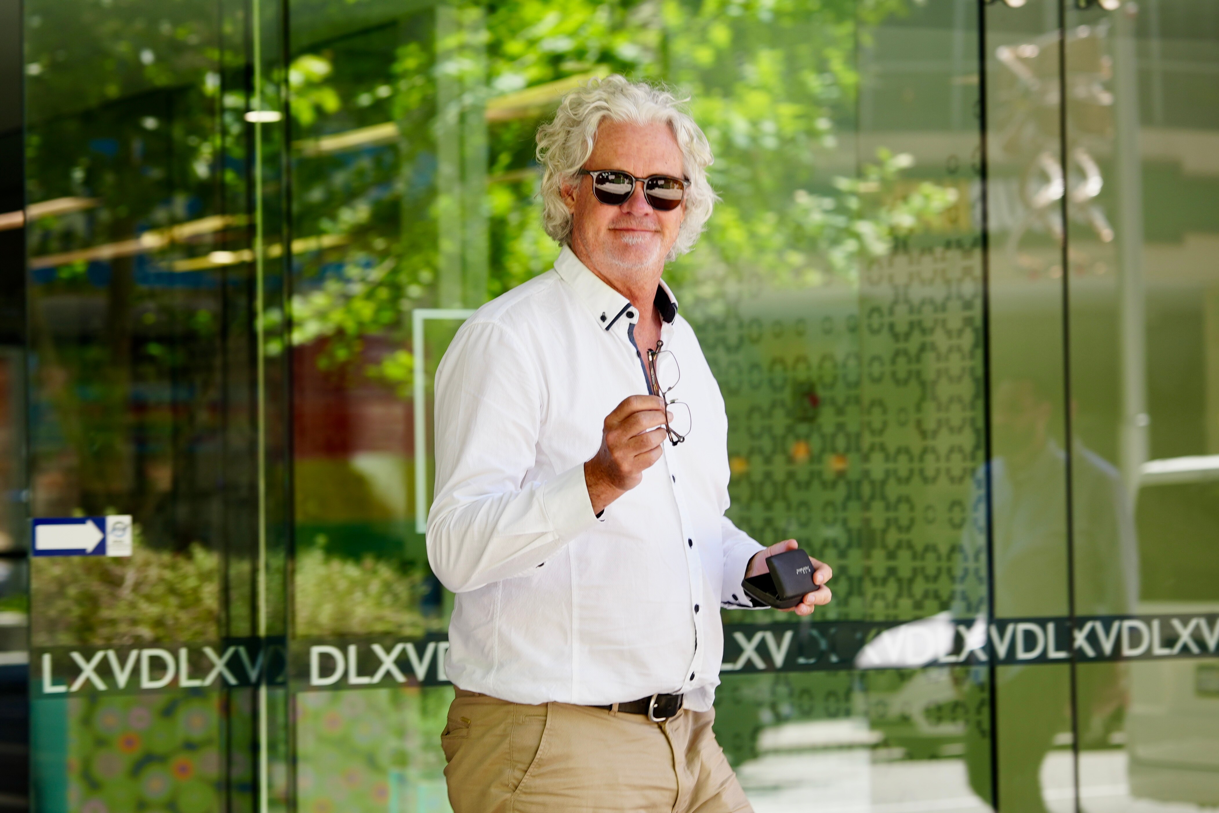 Um homem de cabelos grisalhos e óculos escuros, vestindo uma camisa branca, sorri para a câmera ao sair do tribunal.