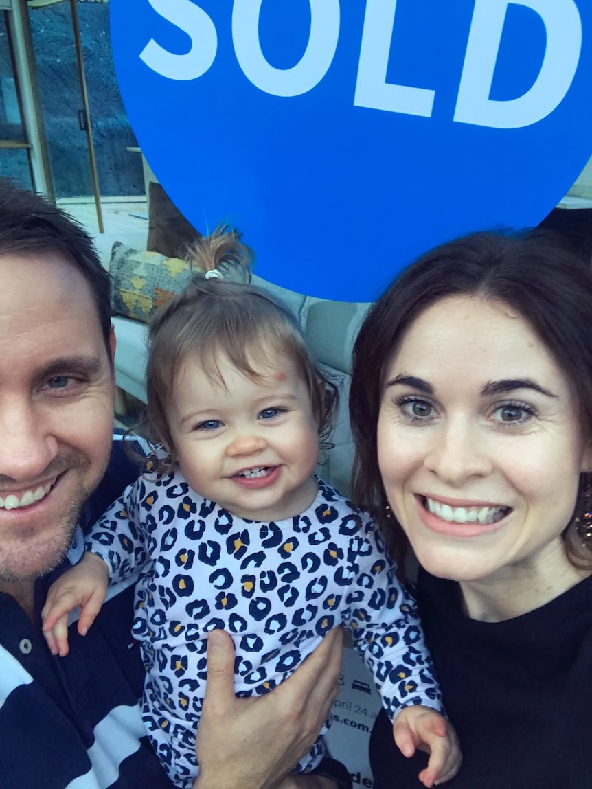 Stuart and Nikki Beard pose with their baby in front of a 'sold sign' for their new house.