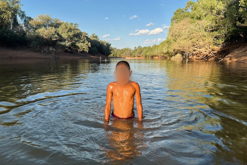 A small boy with a pixelated face stands in a river