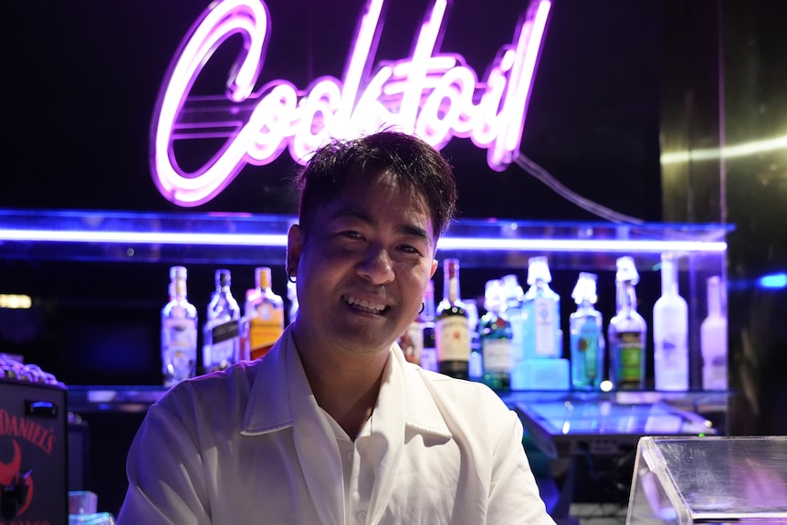 A man with dark hair standing smiling in front of a neon-lit nightclub bar