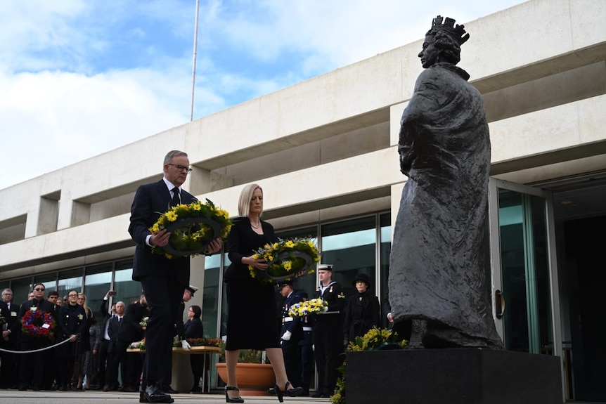 A man in a suit lays a wattle wreath at the base of a statue of the Queen