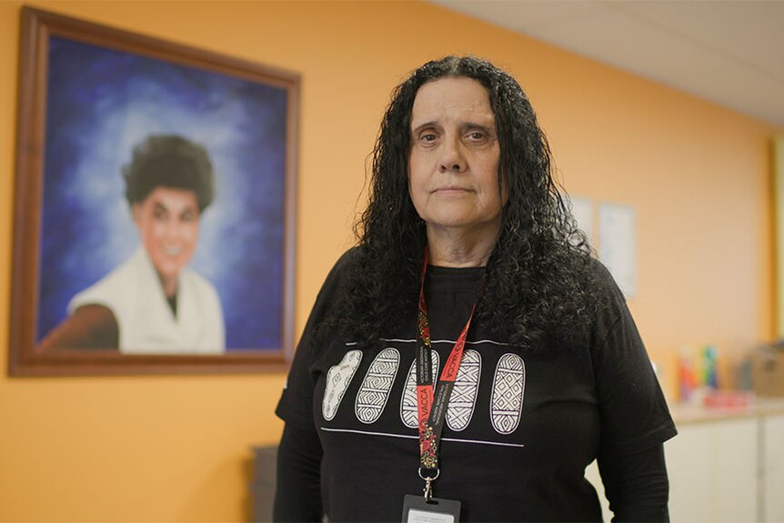 A middle-aged Aboriginal woman wearing a lanyard and a black T-shirt looks at the camera.