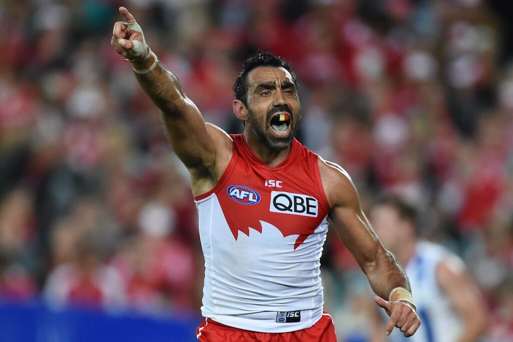 Goodes cheering with one hand in the air pointing, he wears a mouth guard with the Aboriginal flag colours