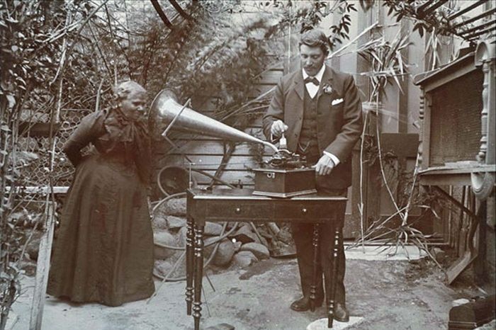 Black and white image of woman and man standing wearing Victorian clothing listening to a gramophone 