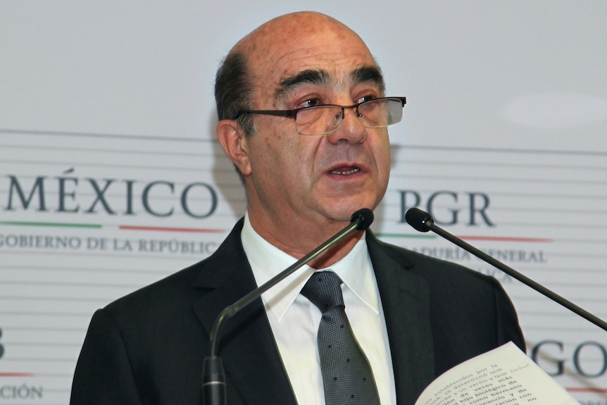 A bald man with bushy eyebrows and glasses stands in front of two microphones