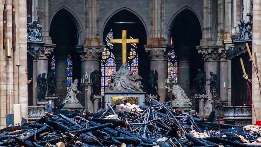 A view of a cross inside Notre Dame with intact statues in the background and burnt and blackened wood in the foreground.