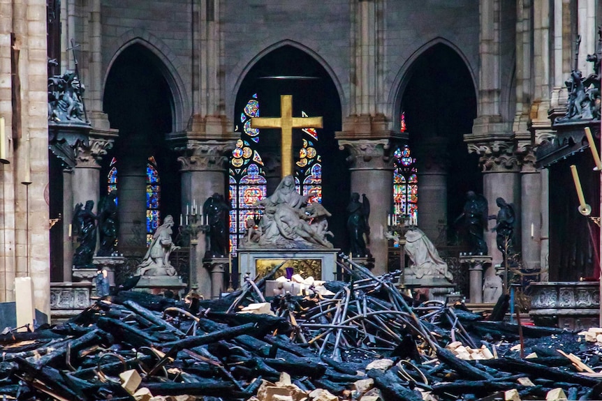 A view of a cross inside Notre Dame with intact statues in the background and burnt and blackened wood in the foreground.