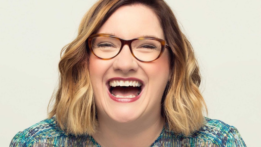 Sarah Millican looks at the camera and laughs in front of a white backdrop.