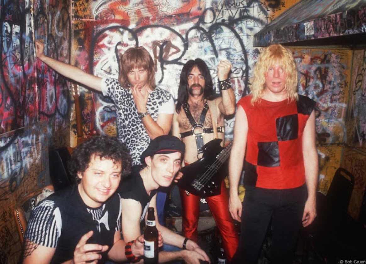 A group of young men dressed in typical 1970s rock star attire stand in a graffitied room.