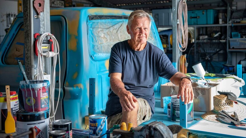 A mid shot of a man sitting on a bench in front of a blue truck in a shed