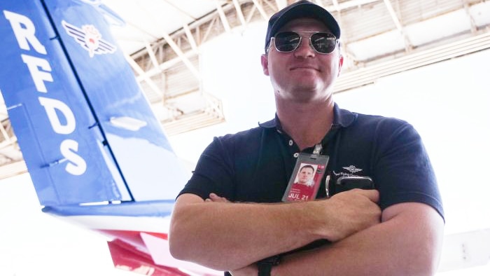 A pilot stands, armed crossed and wearing aviators, before an RFDS plane.