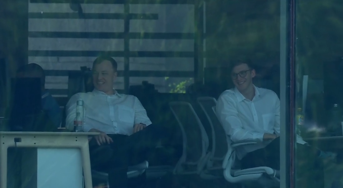 Two men in white office shirts smile in an office while looking out the window.