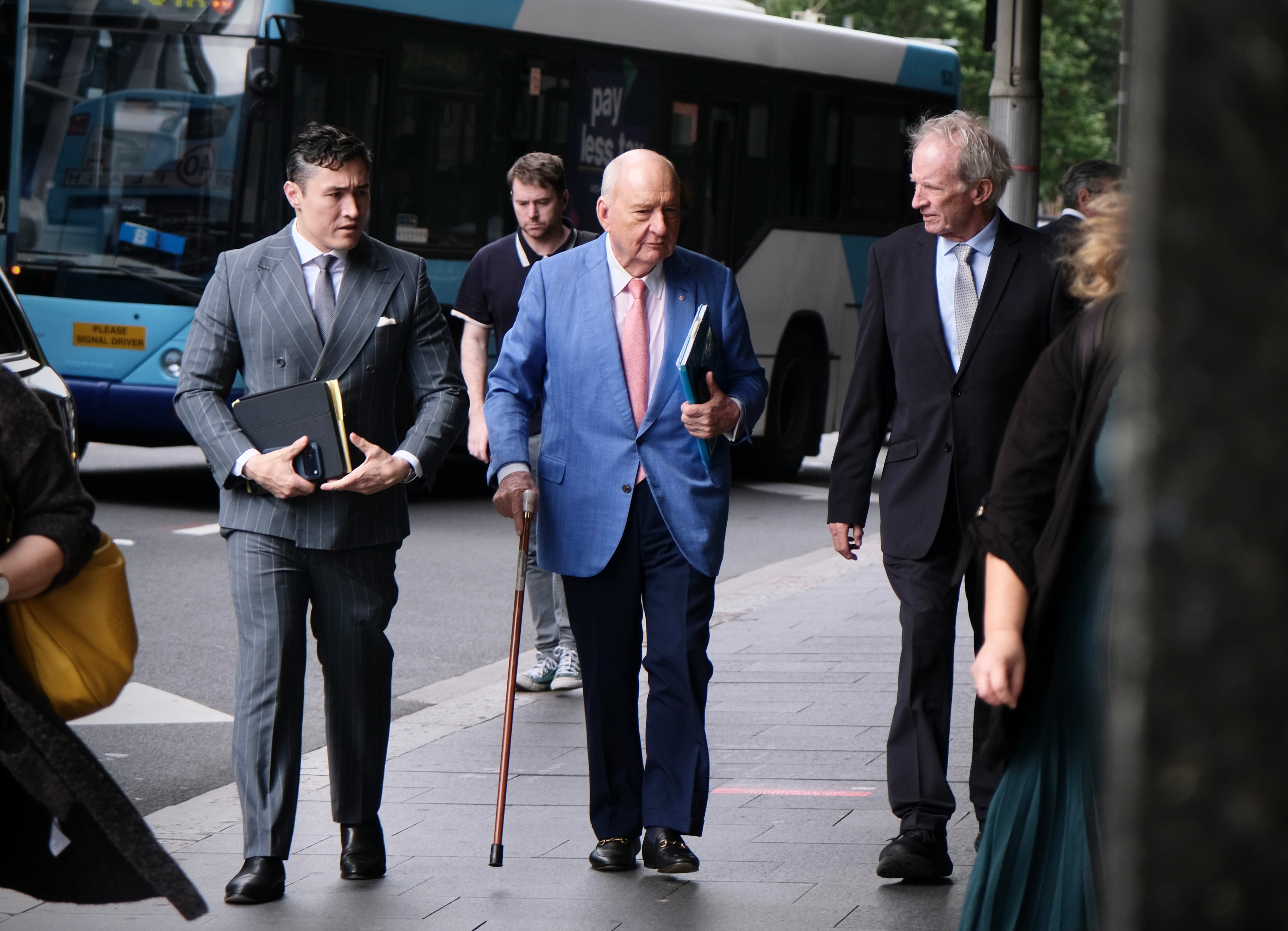 Man in blue jacket, pink tie and walking stick walks with two men in suits either side of him