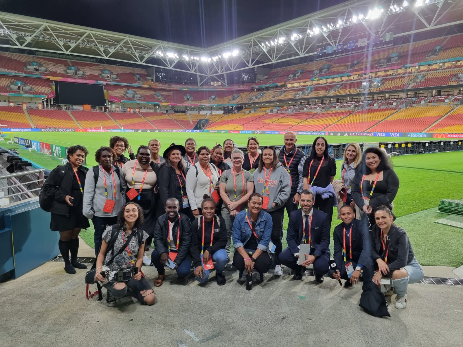 Group of women and some male trainers posing for a photo on the turf of an empty stadium with grandstand in background.