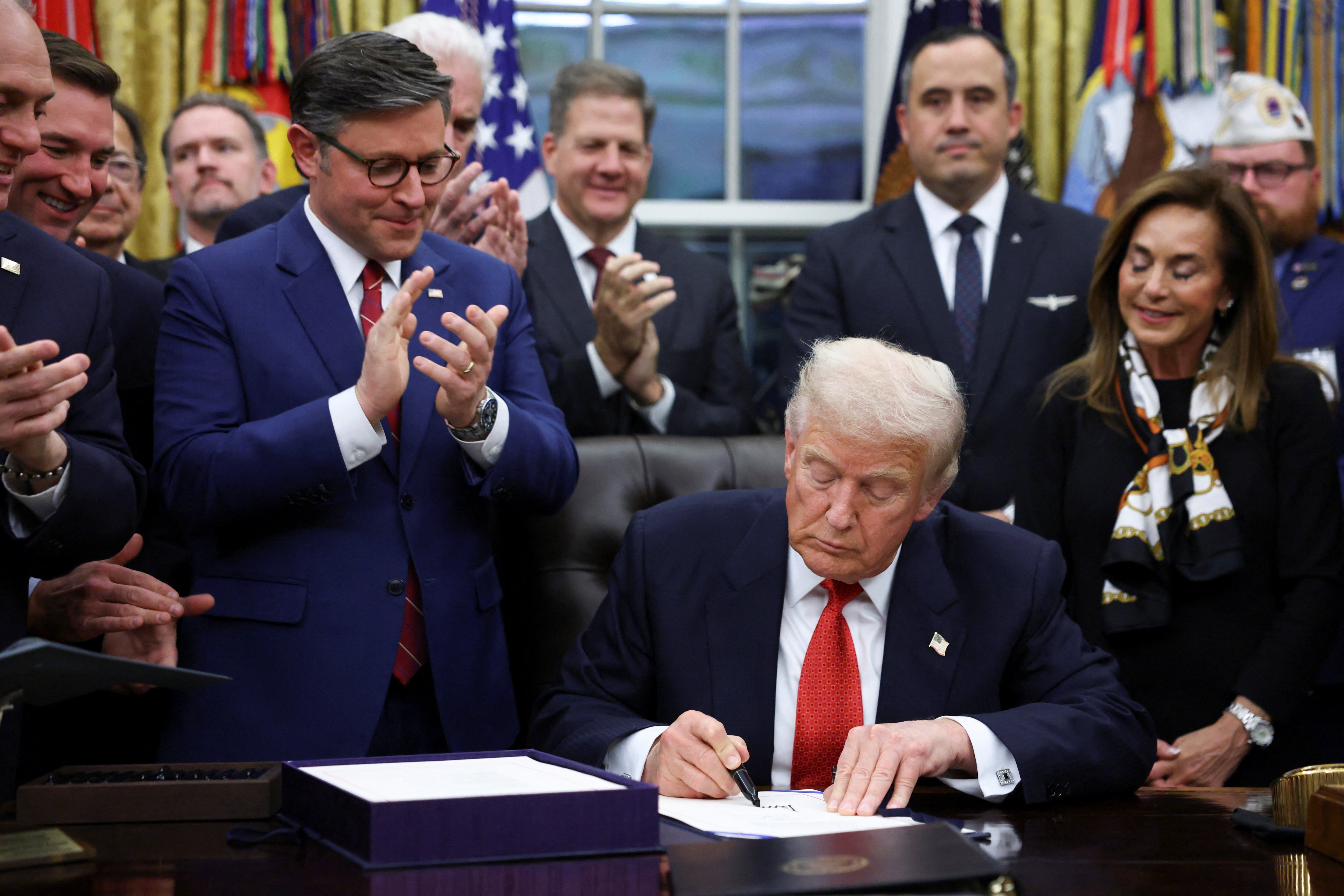 Donald Trump signs a document while a group of people stands around him clapping