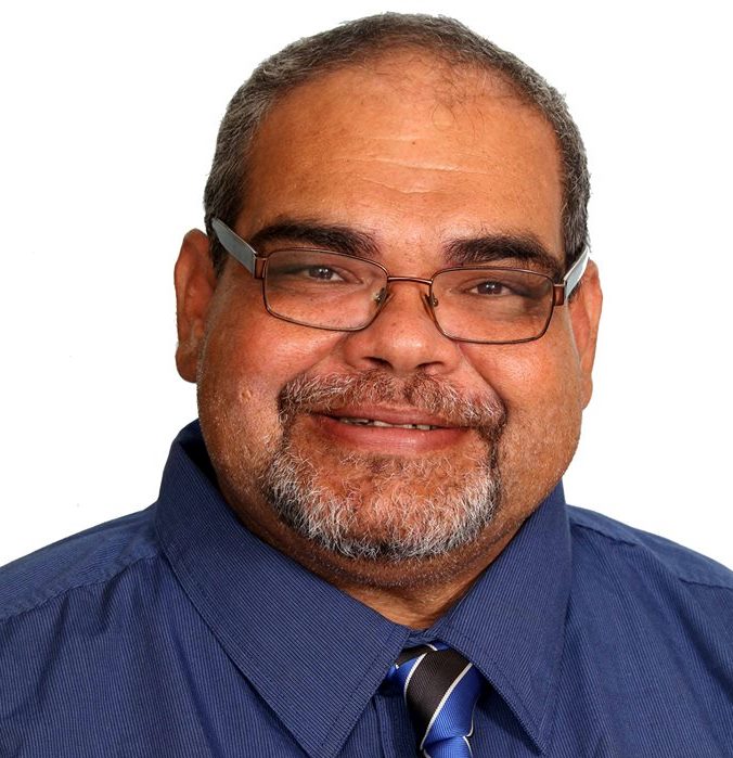 An Indigenous man with shaved hair and glasses smiling, looking at the camera, with blank background