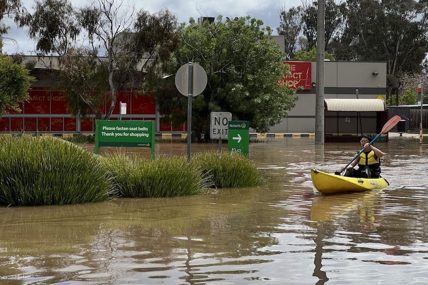 A man canoes through a flooded carpark in a yellow canoe on a sunny day. 